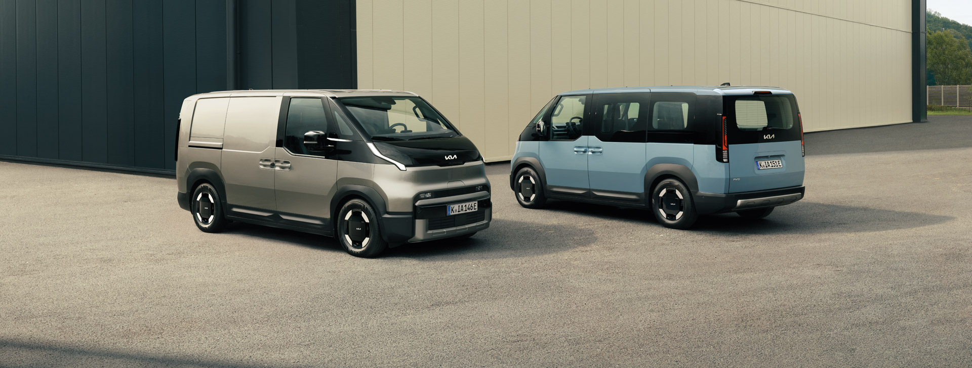 Der Kia PV5 Cargo in schräger Vorderansicht und der Kia PV5 Passenger in schräger Hinteransicht zusammen vor einem gewerblichen Gebäude
